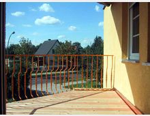 Ein Balkon mit Holzboden und orangefarbenem Metallgeländer, an der Fassade eines gelben Gebäudes. Im Hintergrund sind Bäume, Häuser und eine ruhige Wohnstraße bei sonnigem Wetter zu sehen.