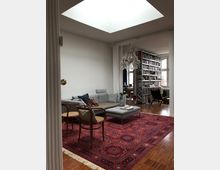 Wohnzimmer in modernem Stil mit Parkettboden und großem roten Orientteppich. Im Vordergrund stehen ein hellgraues Sofa, passende Hocker und Holzstühle, im Hintergrund sind ein Kronleuchter und Regale voller Bücher zu sehen.