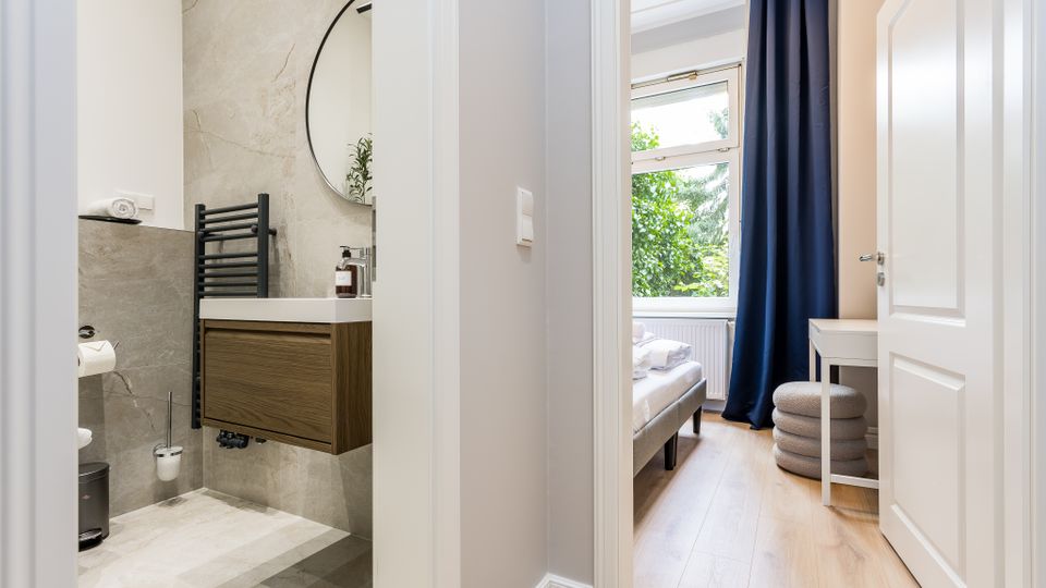 Blick durch einen Flur in ein modernes Badezimmer mit grauen Steinfliesen, wandhängendem Waschtisch und rundem Spiegel sowie in ein angrenzendes Schlafzimmer mit großem Fenster, dunklen Vorhängen und hellem Holzboden. Neben dem Bett stehen ein kleiner Schreibtisch und ein gepolsterter Hocker.