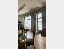 Ein gemütlicher Innenraum mit Holzboden, klassischen Stühlen und einer Tischlampe. Große Fensterfronten lassen Tageslicht einfallen, und draußen sind Teile einer Straße und Baustellenabsperrungen zu sehen.