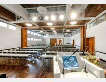 Tagungsraum mit moderner Ausstattung, hellem Holzfußboden und weißen Wänden. Der Raum ist mit langen Tischen und Stühlen in Reihen angeordnet, einer Projektionsfläche und einem Rednerpult mit Mikrofonen im Vordergrund ausgestattet.