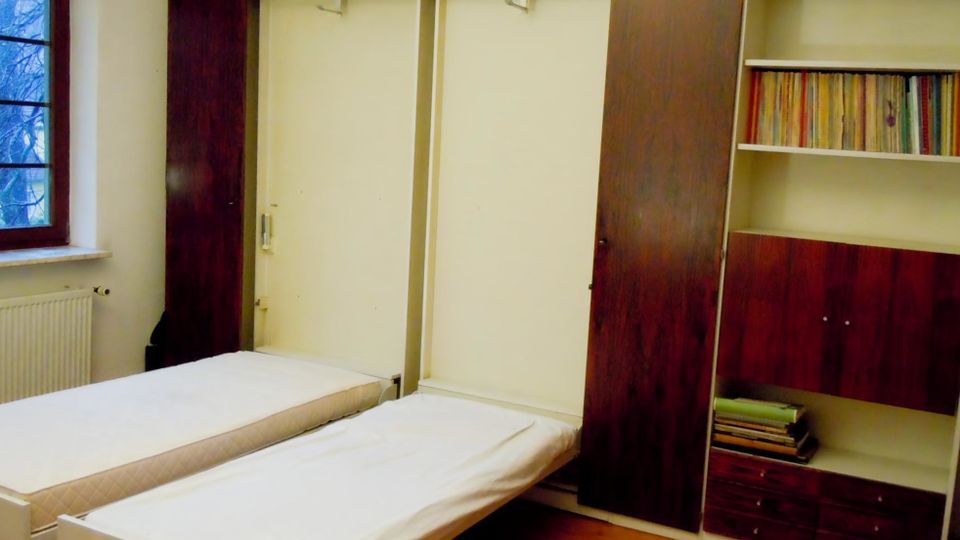 Ein schlichtes Schlafzimmer mit zwei Einzelbetten vor einem großen, wandfüllenden Einbauschrank in dunklem Holz, teils mit offenen Regalen und Schubladen. Links befindet sich ein Fenster mit Blick nach draußen und darunter ein Heizkörper; der Boden ist aus Holz.