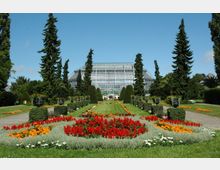 Großer botanischer Garten mit symmetrisch angelegten Blumenbeeten in verschiedenen Farben, flankiert von hohen Nadelbäumen und sorgfältig geschnittenen Sträuchern. Im Hintergrund befindet sich ein großes, gläsernes Gewächshaus mit einer Kuppel.