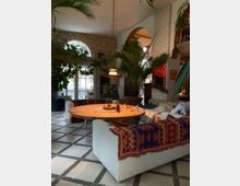 Wohnzimmer mit mediterranem Stil, gekennzeichnet durch Steinwände, große Rundbogenfenster und viele Pflanzen. Zentral steht ein großer runder Holztisch, umgeben von weißen Sofas mit bunten, gemusterten Decken.