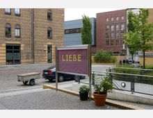 Ein städtischer Platz mit moderner und historischer Architektur, darunter ziegelverkleidete und glatte Gebäudefassaden. Im Vordergrund steht ein Schaukasten mit der Aufschrift „LIEBE“ vor einem Pkw mit Anhänger und mehreren Blumentöpfen.