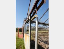 Außenansicht eines verlassenen Gewächshauses mit rostigen Metallrahmen, Glasdächern und einigen offenen Türen. Im Vordergrund eine Metallstange, im Hintergrund trockene Erde und überwucherte Vegetation.