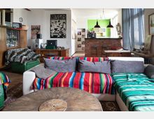 Wohnzimmer in einem urbanen Stil mit einer weißen Couch, bedeckt mit bunten Decken und grauen Kissen, und einem Couchtisch aus Holz. Im Hintergrund eine rote Backstein-Bar mit Barhockern, eine grüne Wand in der Küche, Regale und Poster an den Wänden sowie hohe Fenster mit blauen Vorhängen.