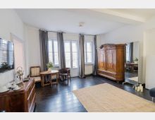 Heller Wohn- oder Hotelzimmerraum mit dunklem Holzboden, großen Fenstern und Balkontür mit langen Vorhängen. Im Raum stehen ein antiker Holzschrank, kleine Sitzgruppe mit Tisch, Kommode mit Tablett sowie ein großer Standspiegel neben einem Teppich.