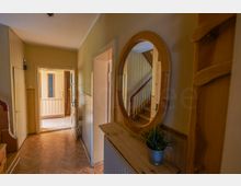Schmaler Flur in einem Wohnhaus mit Holzpaneelen, Fischgrätparkett und einer Kommode mit rundem Wandspiegel. Am Ende führt eine offene Tür in einen helleren Raum, daneben ist ein Treppenaufgang mit Holzgeländer zu sehen.