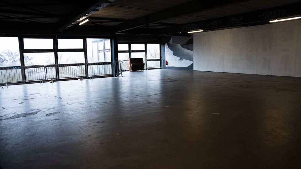 Großer, leerer Innenraum in einer Industriehalle mit dunkler Decke, Betonboden und langen Fensterfronten entlang einer Seite. Metallgeländer stehen vor den Fenstern; an der Stirnseite sind eine breite weiße Wand, eine Treppe und Deckenleuchten zu sehen.