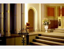 Innenraum einer Kirche mit dekorativen Säulen, die goldene Verzierungen tragen. Der Bereich zeigt eine erhöhte Kanzel, eine Blumenvase auf einem Sockel und einen Kerzenständer; rechts steht ein Altartisch mit Kerze und einer Statue.