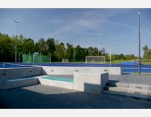 Sportplatz mit blauen Kunststoffbahnen, Basketballkorb, Toren und grünen Netzen für Wurfsportarten, umgeben von Bäumen. Im Vordergrund befinden sich graue Betonstufen und Sitzflächen.