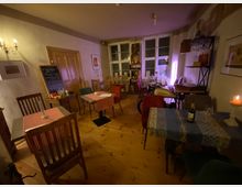 Gemütlicher Gastraum eines kleinen Cafés mit Holzdielenboden, mehreren kleinen Tischen mit gemusterten Tischdecken und gemischten Stühlen. Hinten stehen hohe Fenster, ein Schrankregal und eine Kreidetafel; an der Wand sorgen Kerzen und eine indirekte Lichtquelle für warmes Licht.