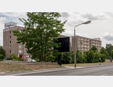 Mehrstöckiges Wohngebäude aus Beton mit einem Aldi-Logo auf dem Dach. Im Vordergrund befinden sich eine kleine Grünfläche mit Bäumen, ein leeres Werbeschild, eine Straßenlaterne und ein Gehweg entlang einer Straße.