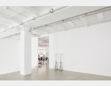 Weißer, minimalistischer Innenraum eines Studios oder Lofts mit sichtbaren Deckenrohren und -verkabelungen. Im Hintergrund befindet sich ein Raum mit großen Fenstern, Tisch und Stühlen, während im Vordergrund zwei zusammengeklappte Metallstative an der Wand lehnen.