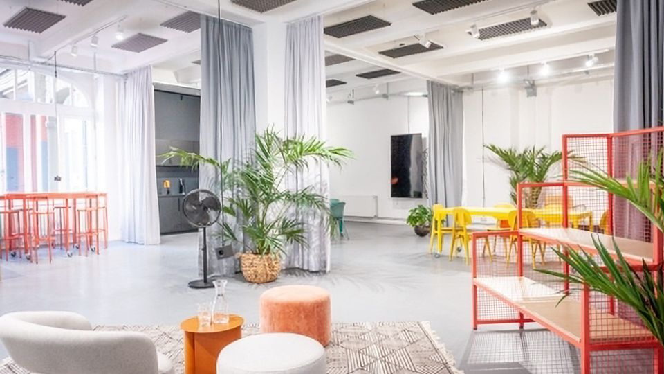 Heller, offener Studio- oder Coworking-Raum mit hohen Decken, Akustikpaneelen, großen Fenstern und grauen Vorhängen als Raumteiler. Im Vordergrund stehen Loungesessel und Hocker auf einem Teppich, daneben Pflanzen; im Hintergrund sind gelbe Tische und Stühle sowie rote Gitterregale und Stehtische zu sehen.