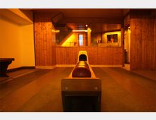 Holzverkleidete Kegelbahn in einem Innenraum mit warmem Licht, sichtbaren Kugeln auf der Bahn und einer Fensterfront, die einen Blick in einen angrenzenden Raum ermöglicht.