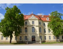 Barockes Gebäude mit heller Fassade und roten Ziegeldächern, verziert mit Stuckelementen über den Fenstern. Im Vordergrund stehen zwei große Bäume und ein Weg führt zu einem Haupteingang mit Treppe.