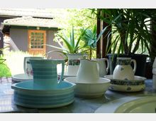 Keramikkrüge und Schüsseln stehen auf einer Arbeitsfläche am Fenster, dahinter sind Topfpflanzen und ein Garten mit einem Gebäude mit Holztür zu sehen. Die Szene wirkt ländlich und zeigt Details eines sonnigen Außenbereichs.