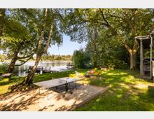 Ufernaher Garten am See mit schattenspendenden Bäumen, Steg am Wasser und Rasenfläche. Im Vordergrund stehen eine Tischtennisplatte auf einer Terrasse sowie mehrere bunte Gartenstühle; rechts ist ein überdachter Gebäudeteil zu sehen.