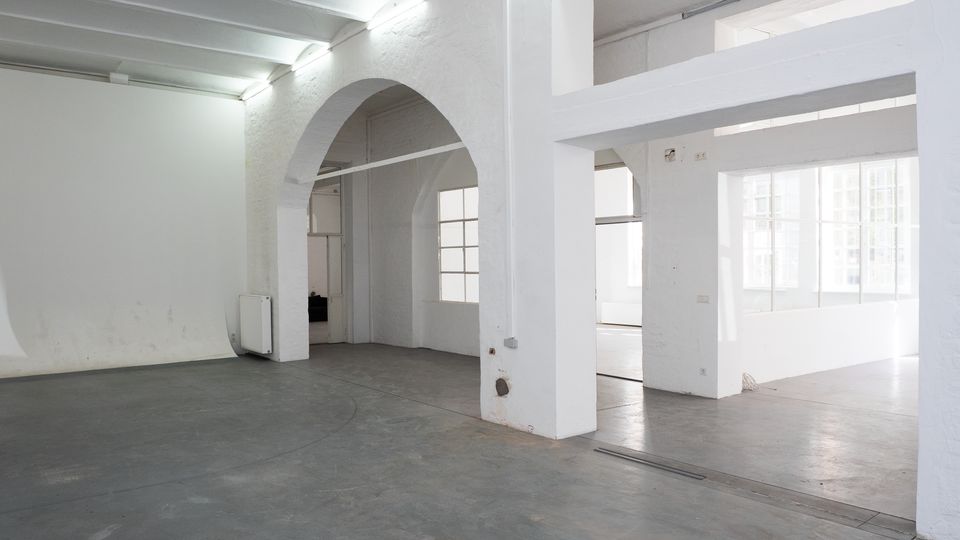 Heller, leerer Innenraum im Loft- oder Industrie-Stil mit weiß gestrichenen Wänden, Sichtdeckenbalken und grauem Betonboden. Große Rundbogenöffnung, breite Stützpfeiler und hohe Fenster lassen viel Tageslicht in den Raum.