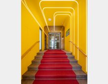 Ein gelb gestrichener Eingangsbereich mit einer Treppe, die mit rotem Teppich belegt ist. An den Seiten der Wände befinden sich Holzhandläufe, und weiße, leuchtende LED-Streifen formen geometrische Muster an Decke und Wänden. Oben führt eine Glastür zu einem weiteren Raum.