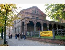 Backsteingebäude mit Rundbögen und Arkadengang, umgeben von einem Metallzaun mit niedriger Backsteinmauer; vor dem Gebäude stehen mehrere Personen, und ein gelbes Banner mit der Aufschrift „Chorkonzert Sonntag 18 Uhr“ hängt am Zaun. Im Vordergrund stehen Bäume, der Gehweg ist mit herbstlichem Laub bedeckt.