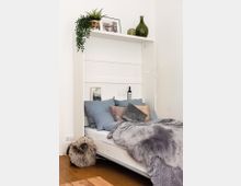 Kompakter Schlafbereich mit einem hochklappbaren weißen Wandbett, das mit blauer und grauer Bettwäsche sowie Kissen dekoriert ist. Oberhalb des Bettes befinden sich Pflanzen, gerahmte Bilder und grüne Glasvasen, der Boden ist aus Holz, und es gibt eine flauschige Sitzauflage in der Nähe.