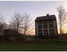 Zweistöckiges, verlassenes Gebäude mit zerbrochenen Fenstern und verwitterter Fassade, umgeben von kahlen Bäumen und Wiesen. Im Hintergrund ein weiterer, teils verdeckter Ziegelbau unter einem farblich dezenten Abendhimmel.