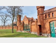 Backsteinmauer und Tor eines historischen Gebäudekomplexes mit neogotischen Elementen, bestehend aus Türmen, Rundbögen und spitz zulaufenden Giebeln. Im Hintergrund steht ein rotes Backsteinbauwerk mit großen Fenstern und dekorativen Verzierungen, umgeben von Bäumen und einer Wiese.