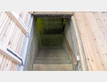 Holztreppe, die in einen einfachen Kellerraum führt, umgeben von Betonwänden und seitlich angebrachten Holzgeländern. Der Keller hat einen betonierten Boden und eine dunkle, schlichte Umgebung.