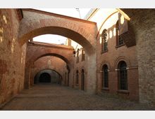 Backsteinbau mit einem gepflasterten Innenhof, umgeben von hohen Ziegelwänden mit Fensterbögen und mehreren steinernen Rundbögen, die die Gasse überspannen. Im Hintergrund führt ein dunkler Durchgang in einen weiteren Bereich.