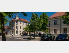 Straßenecke in einem kleinen Ortszentrum mit mehrstöckigen, hell verputzten Altbauten und roten Ziegeldächern, darunter ein Eckhaus mit kleinem Turmaufsatz. Entlang der Straße stehen parkende Autos, mehrere Verkehrsschilder und dichte Bäume, die Teile der Fassaden und den Gehweg säumen.