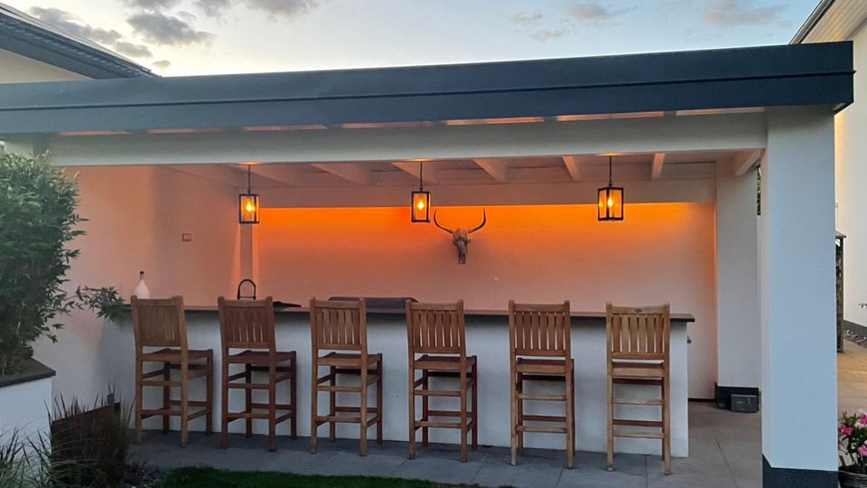 Überdachte Außenbar im Garten mit weißer Rückwand, dunkler Theke und sechs hohen Holz-Barhockern. Unter dem Dach hängen drei Laternenleuchten, und hinter der Theke sorgt eine orangefarbene indirekte Beleuchtung mit dekorativem Longhorn-Schädel an der Wand für Akzente.