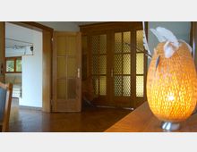 Holzinterieur eines Wohnraums mit Glastüren, die ein Wabenmuster aufweisen, und einer geöffneten Tür, die in einen anderen Raum mit Sofa und Fenstern führt; im Vordergrund eine leuchtende, geflochtene Lampe auf einem Tisch.