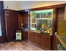 Innenraum mit dunkler Holzvertäfelung und eingebauten Vitrinen, in denen verschiedene Ausstellungsobjekte und Poster hinter Glas präsentiert werden. Vor der Vitrine stehen ein kleines Retro-Heizgerät und auf einer Nische sind dekorative Geräte wie eine alte Nähmaschine und ein Samowar zu sehen.