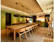 Moderner Speise- oder Gemeinschaftsraum mit langem Holztisch und vielen Stühlen, darüber mehrere schwarze Pendelleuchten an einer Holzdecke. Grüne Akzentwand, offene Regale mit Beleuchtung sowie ein großer Holzständer neben bodentiefen Fenstern und Holzboden.