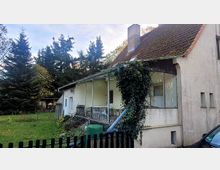 Ein kleines, älteres Wohnhaus mit verputzter Fassade und einem mit Ziegeln gedeckten Satteldach, teilweise bewachsen von Efeu. Im Vordergrund befinden sich ein Garten mit einer Rasenfläche, ein Zaun und ein grüner Regenwassertank, dahinter stehen hohe Nadelbäume.