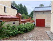 Gepflasterter Hof mit einer hellen Hauswand auf der linken Seite und einer roten Backsteinmauer mit Ziegelabdeckung, die den Bereich begrenzt. Im Hintergrund steht ein Gebäude mit einer großen roten Holztür und einem Dach aus Metallblech, umgeben von grüner Vegetation.