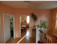 Ein Wohnbereich in warmen Pastelltönen mit Holzboden, einem Geländer rechts und drei Türen, die in andere Räume führen. An den Wänden hängen Bilder, und es stehen mehrere Zimmerpflanzen und eine Holzkommode im Raum.