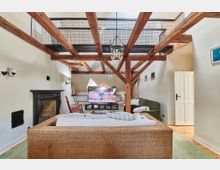 Wohnzimmer mit sichtbaren Holzbalken und einer hohen Decke, ausgestattet mit einem Kamin, einem großen Flachbildfernseher, einem geflochtenen Sofa, einem grünen Sofa und Holzboden. Eine offene Empore und weiße Wände mit minimaler Dekoration runden den Raum ab.