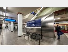 U-Bahn-Station Potsdamer Platz in Berlin mit Sitzbänken, einem Informationsschild und Hinweisschildern zu Ausgängen. Im Hintergrund sind Rolltreppen, Säulen, und eine teilweise sichtbare S-Bahn zu sehen.