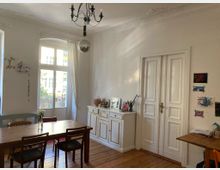 Helles Wohnzimmer mit Holzboden und klassischer Einrichtung. Zu sehen sind ein großer Holztisch mit Stühlen, eine weiße Kommode mit Dekorationsgegenständen sowie eine Doppelflügeltür und ein großes Fenster, durch das Tageslicht einfällt.