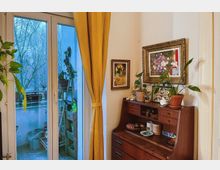 Wohnraum mit einer gläsernen Balkontür, die zum Außenbereich mit Pflanzen und Gartenutensilien führt. Im Raum steht ein antiker Sekretär mit Dekoobjekten und Topfpflanzen, daneben hängen gerahmte Bilder an einer hellen Wand, ergänzt durch einen gelben Vorhang.