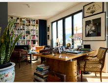 Helles Wohnzimmer mit einer großen Bücherwand, einem Schreibtisch aus Holz und verschiedenen Sitzgelegenheiten. Große Fenster lassen viel Tageslicht herein, und draußen ist eine Terrasse mit Sonnenschirm sichtbar; an den Wänden hängen gerahmte Kunstwerke.