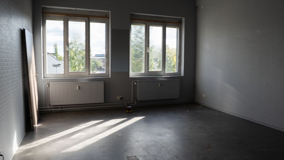 Leerer, grauer Innenraum mit zwei großen Fenstern an der Stirnwand, darunter Heizkörper und sichtbaren Leitungen. Glatte Betonbodenfläche mit Lichtstreifen durch die Fenster und einer an die linke Wand gelehnten Türplatte.