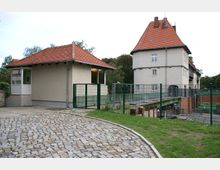 Zwei Gebäude mit hellen Putzfassaden und roten Ziegeldächern, eines mit einem steilen Turmdach; im Vordergrund ein gepflasterter Weg, im Hintergrund ein Zaun und Grünflächen.