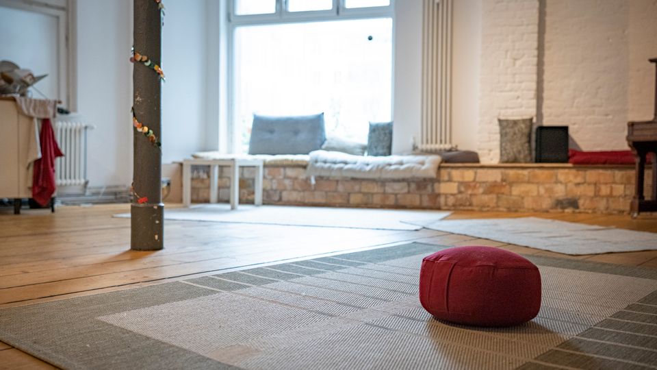 Heller Raum mit Holzfußboden und großen Teppichen, in der Mitte ein rotes Sitzkissen. Im Hintergrund eine niedrige Sitzbank mit Kissen vor einem großen Fenster, weiße Ziegelwand und ein Heizkörper.