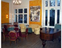 Ein eleganter Raum mit hellgelben Wänden und großen Fenstern, eingerichtet mit Polstermöbeln, kleinen Tischen mit Tischdecken und einem Mahagoni-Flügel im Vordergrund. An der Wand hängt ein großformatiges Kunstwerk, und eine Deckenleuchte sowie Wandleuchten sorgen für stimmungsvolle Beleuchtung.