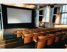 Ein kleiner Kinosaal mit Holzstühlen in Reihen, einem großen, weißen Kinobildschirm und industriellen Elementen wie Betonwänden und hohen Fenstern, die Tageslicht hineinlassen.